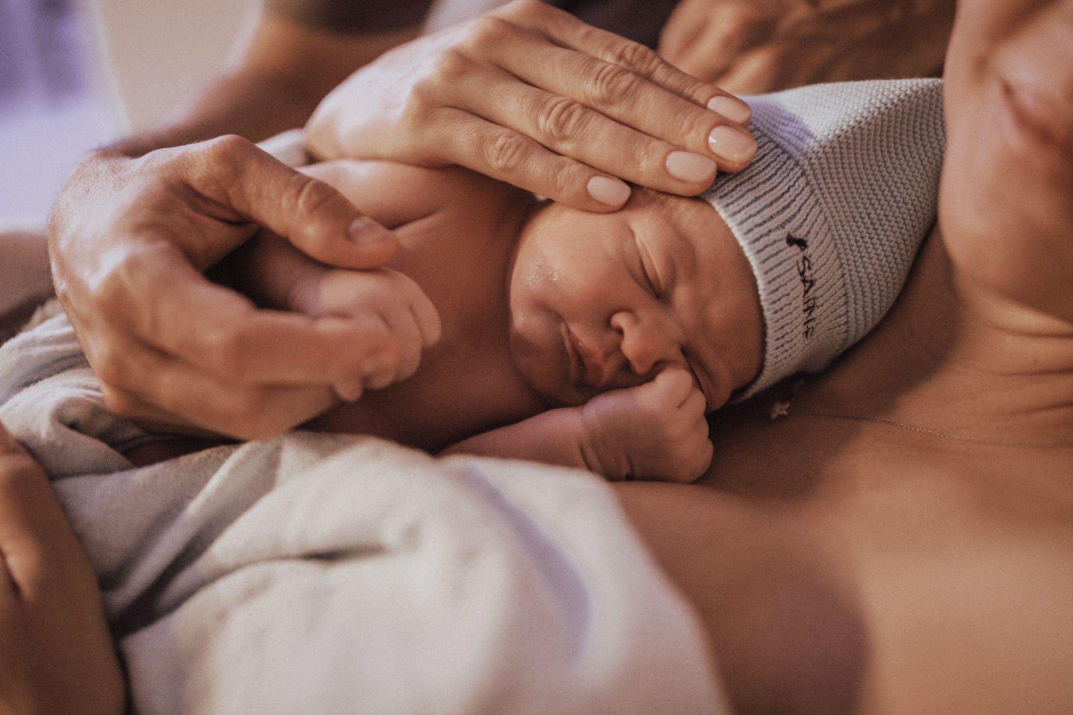 Recém-nascido dormindo de touquinha do Saine no colo da mãe, com a mão dela acariciando a cabeça
